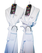 Two white textured bathrobes hanging on a rack, each with a black hangtag and a matching pocket, photographed against a light wall from Hotel ZAZZ.
